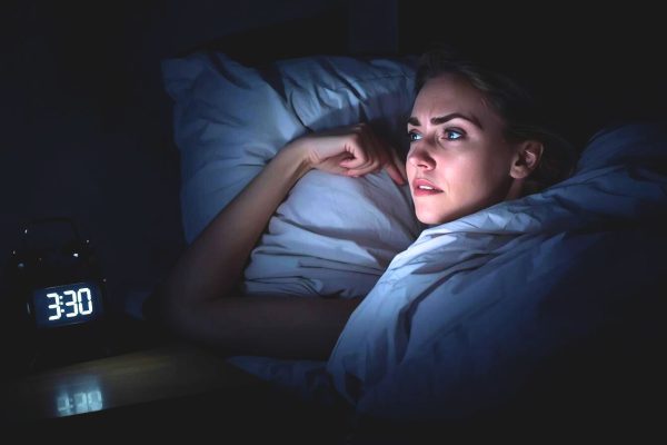 Молодая женщина лежит в постели в темноте и с грустным выражением лица смотрит на светящийся циферблат электронного будильника, показывающего 3:30 ночи.